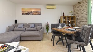 A living room with a grey sofa, a dining table set for four, and a painting of a sunset on the wall. - Apartment 2333/111878 (Istria - Pula), 3000m from the beach (Pula)