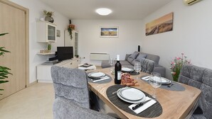 A modern dining area with a wooden table set for four, surrounded by grey upholstered chairs. A television is mounted on the wall, and there are decorative plants and artwork on shelves. - Apartment 2333/111878 (Istria - Pula), 3000m from the beach (Pula)