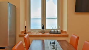 Un comedor con una mesa y sillas, un ventanal con vistas al mar, un refrigerador y una estantería con objetos decorativos.