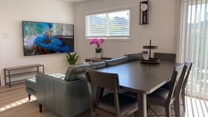 Un comedor moderno con una mesa grande, asientos acolchados, un televisor y una ventana con persianas.