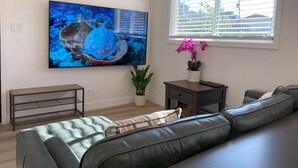 Una sala de estar con un televisor de pantalla plana grande, un sofá, una mesita con una planta en maceta y una ventana con persianas.
