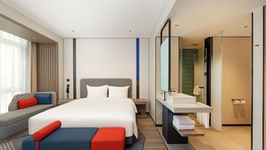 Una camera d'albergo moderna con un letto grande, una poltrona, una panca, un bagno con lavandino e specchio, e vista sulla città.