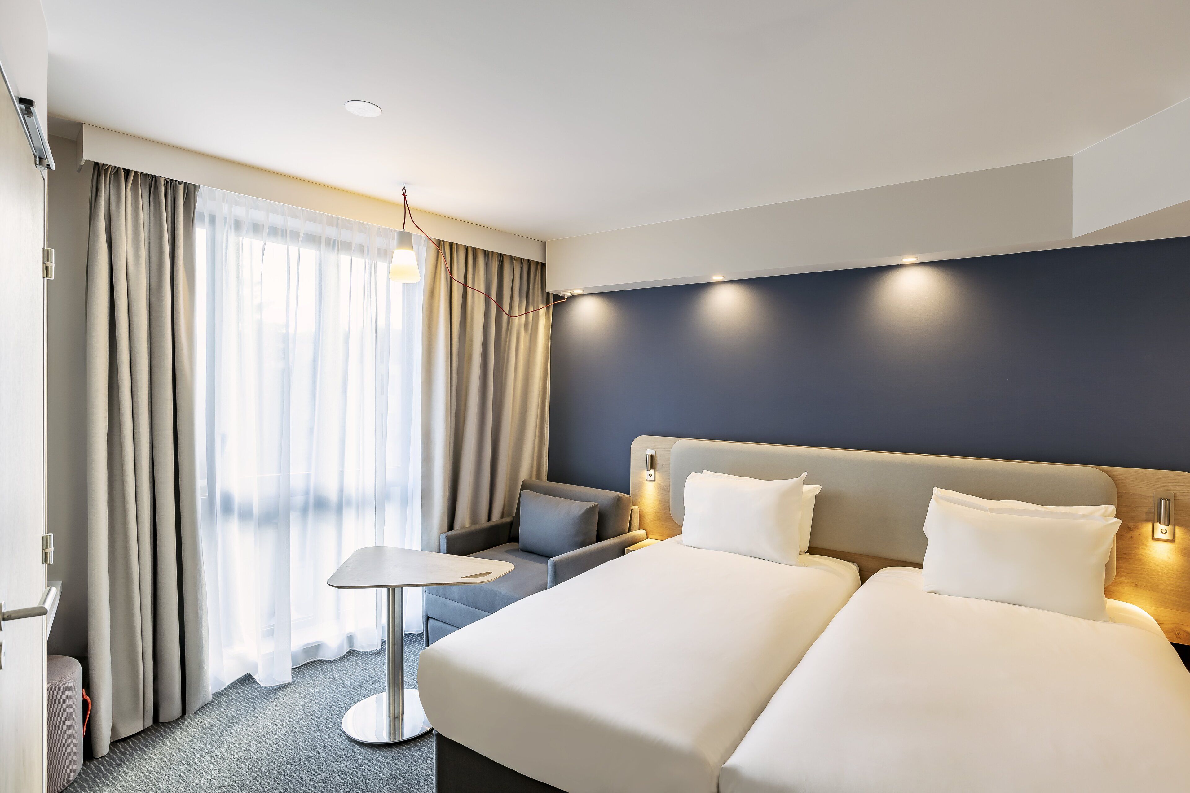 Habitación de hotel con una cama grande, una mesita pequeña, un sofá y vistas a la ciudad a través de cortinas translúcidas.