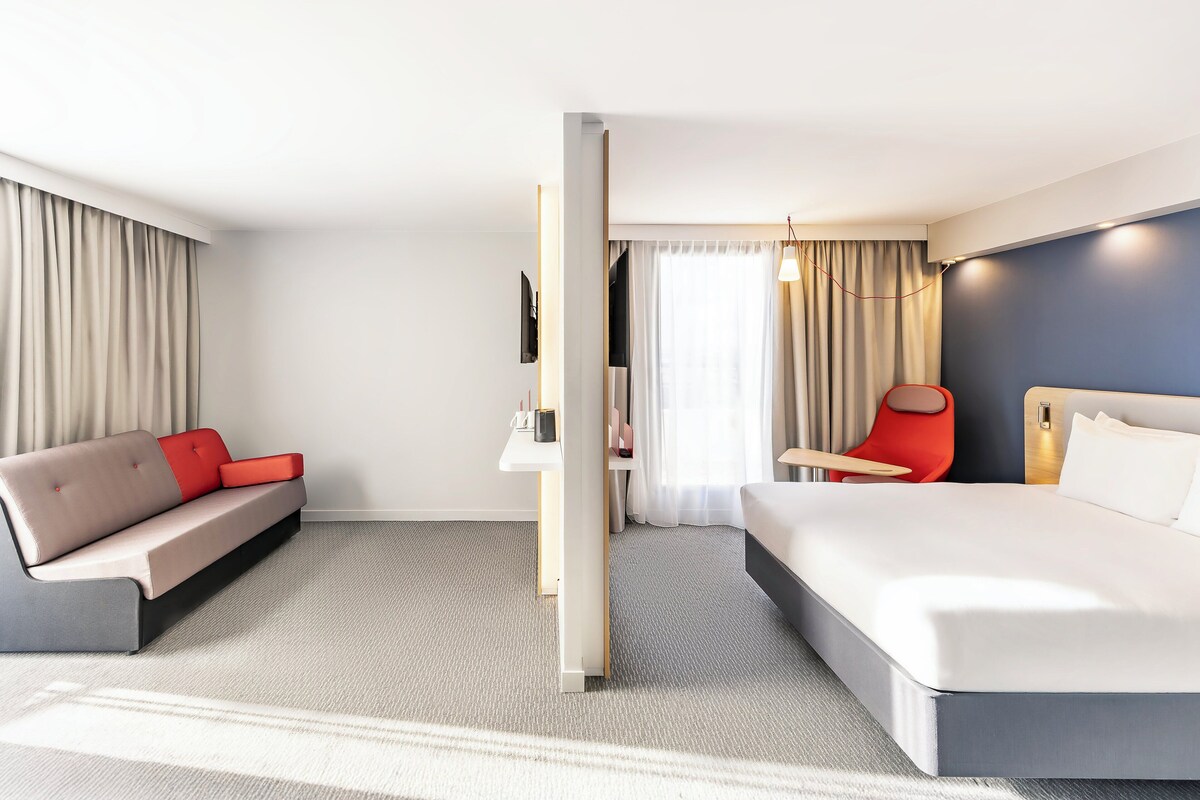 A modern hotel room with a large bed, a sofa, a desk, and a chair.
