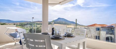 Un balcón con mesa lista para comer, sillas de plástico blanco y vistas a un paisaje montañoso.