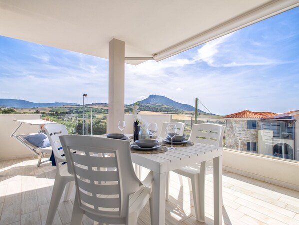 A balcony with a table set for dining, white plastic chairs, and a view of a mountainous landscape.