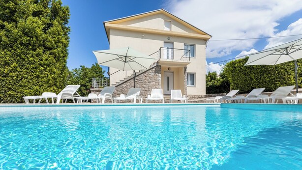 A poolside view of a two-story building with a stone facade, white lounge chairs, and umbrellas.