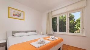 A double bed with an orange blanket, white pillows, and two rolled towels. There is a framed picture on the wall, a bedside table, and a window with curtains.