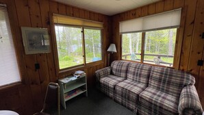 L'intérieur d'une cabane est aménagé avec un canapé à carreaux, un fauteuil, une petite bibliothèque et de grandes fenêtres qui offrent une vue sur l'extérieur.