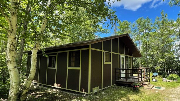 Une petite cabane dotée d’un porche, entourée d’arbres et d’une zone herbeuse.