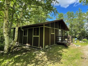 Une petite cabane dotée d’un porche, entourée d’arbres et d’une zone herbeuse.