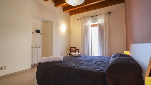 Een slaapkamer met een groot bed, houten balken in het plafond, een radiator, een stoel en een raam met gordijnen.