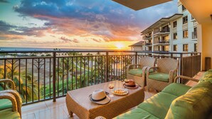 A balcony with wicker furniture, a table with a hat and wine glass, and a view of the ocean and palm trees at sunset.