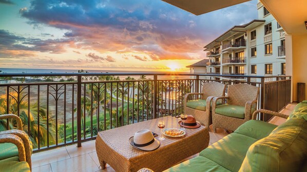 A balcony with wicker furniture, a table with a hat and wine glass, and a view of the ocean and palm trees at sunset. - End of year specials! - BT701 Beautiful 3 Bed, 3 Bath (Kapolei)