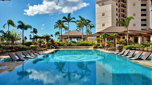 A swimming pool with lounge chairs, palm trees, and a multi-story building in the background.