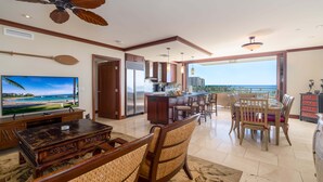 A spacious living area with a large TV, wooden furniture, a ceiling fan, and a view of the ocean.