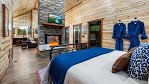 A log cabin-style room with a stone fireplace, a bed with blue bedding, and a view of the kitchen in the background.