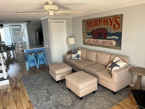 A living room with a beige sectional sofa, two matching ottomans, a grey patterned rug, a ceiling fan, and a wall-mounted sign.