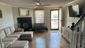 A living room with a sofa, ottoman, fireplace, ceiling fan, and a TV mounted on the wall. - Fenwick Island, 1 block to beach, 2 BR condo, Sleeps 6, 2 parking spots (Fenwick Island)