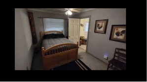 Une chambre avec un lit en bois, une fenêtre avec des stores, un ventilateur de plafond et une porte donnant sur une autre pièce.
