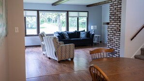 Un salon doté d’un plancher en bois, d’un canapé de couleur foncée, de fauteuils à motifs, d’une table à manger avec des chaises et d’un mur d’accent en briques.