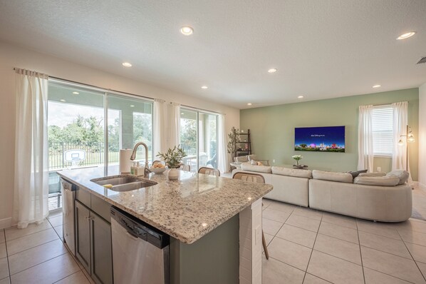 A modern kitchen with a large island, a dining area with a sofa, and a TV mounted on the wall. - Luxury 5BR Villa, Private Pool/Spa, 15 Min Disney! (Kissimmee)