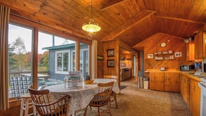Interior acogedor de una cabaña con comedor, techo de madera y ventanas amplias que dan a la naturaleza.