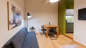 Un espacio habitable pequeño con un sofá, una mesa de comedor con sillas, una lámpara de pie y una cocina visible a través de un vano abierto.