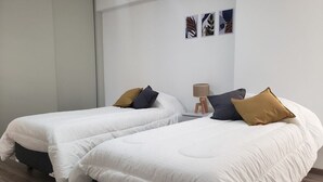 Ein Hotelzimmer mit zwei Betten, jeweils mit weißer Bettwäsche und einer Auswahl an Kissen. Zwischen den Betten steht ein kleiner Tisch mit einer Lampe.