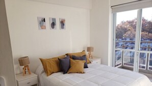 Ein Schlafzimmer mit einem großen Bett, zwei Nachttischen, einem Fenster mit Blick auf Gebäude und drei gerahmten Bildern an der Wand.