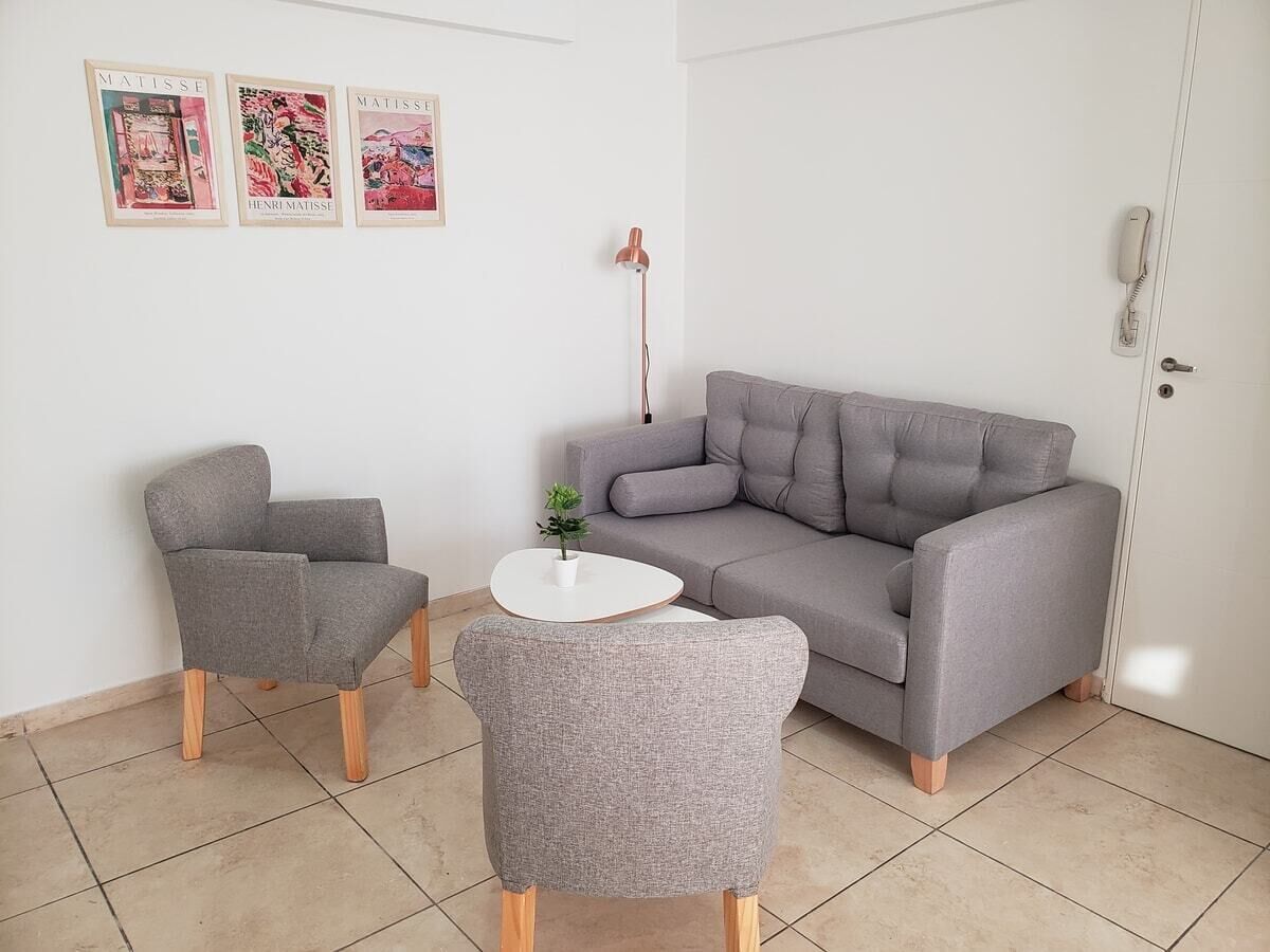 A living room with a grey sofa, two matching armchairs, a small white table, and a potted plant. There are three framed artworks on the wall.