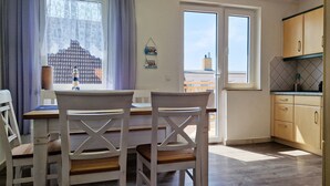 A dining area with two white chairs and a table, a kitchen with wooden cabinets, and a view of buildings through a window. - Weitsicht, Deutschland (Kühlungsborn)