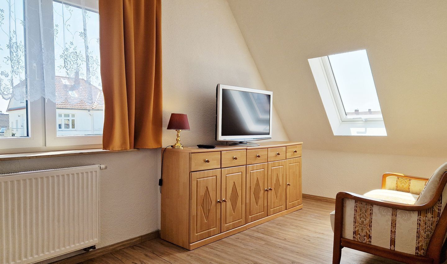 Ein Zimmer mit einem Holzschrank, einem Fernseher darauf, einem Sessel und einem Fenster mit Vorhängen.