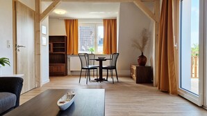 A room with a dining table and chairs, a sofa, a wooden floor, and a view through a window with curtains. - Weitsicht, Deutschland (Kühlungsborn)