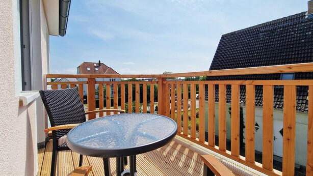 A balcony with a round glass table and two chairs, wooden railings, and a view of neighboring buildings. - Weitsicht, Deutschland (Kühlungsborn)