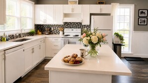 A modern kitchen with white cabinetry, a central island with baked goods and flowers, and a stove. - Downtown Picton Retreat |Near Sandbanks & Wineries (Prince Edward)