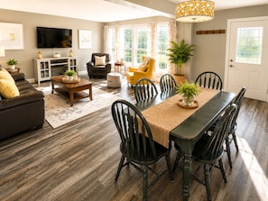 A living room with a dark wooden dining table, black chairs, a sofa, a television, and a large window with curtains. - Downtown Picton Retreat |Near Sandbanks & Wineries (Prince Edward)