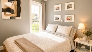 A bedroom with a bed, a nightstand with a lamp, framed pictures on the wall, and a window with curtains. - Downtown Picton Retreat |Near Sandbanks & Wineries (Prince Edward)