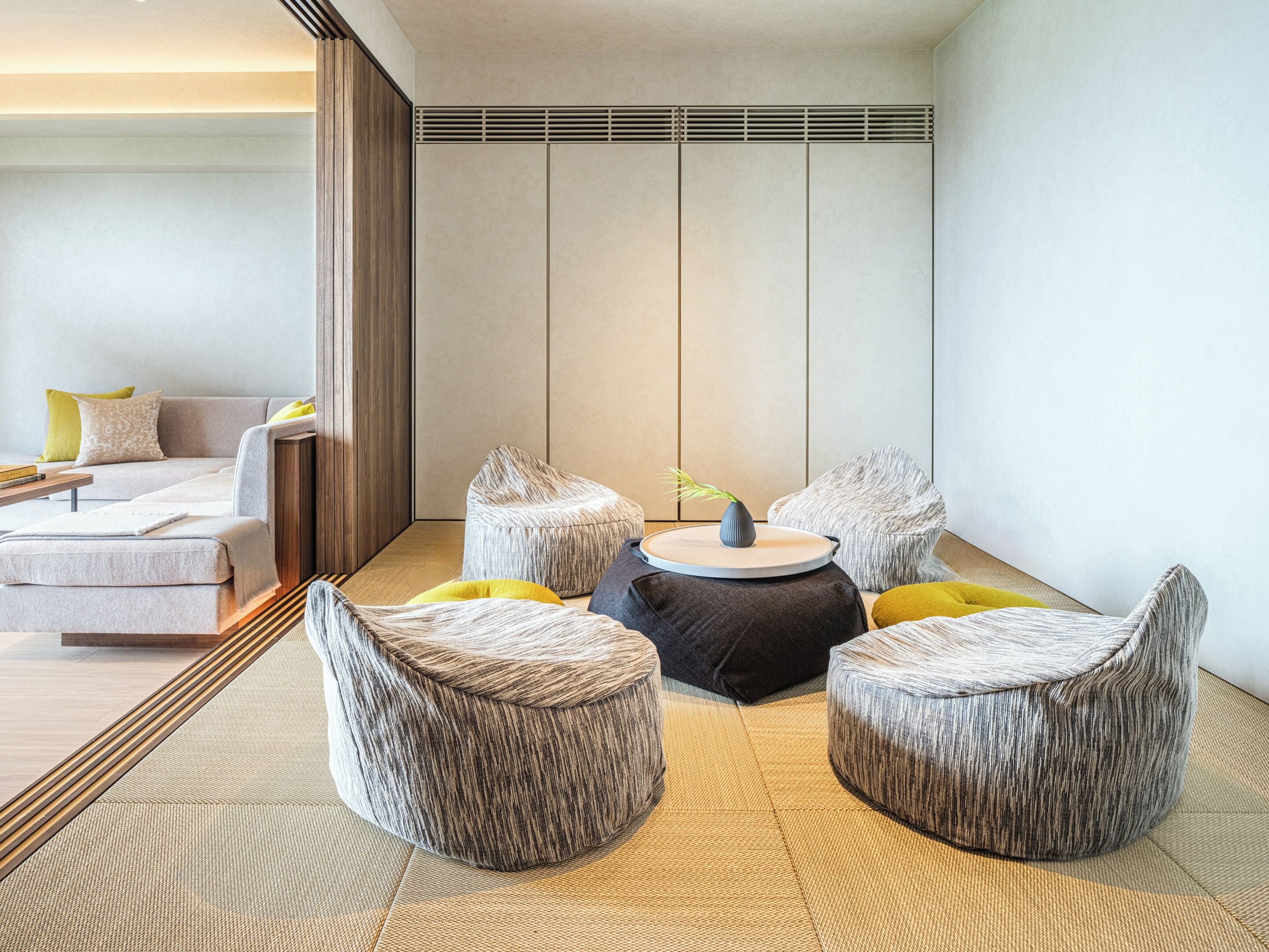 A modern hotel room with a sofa, two round ottomans, and a small table with a vase.