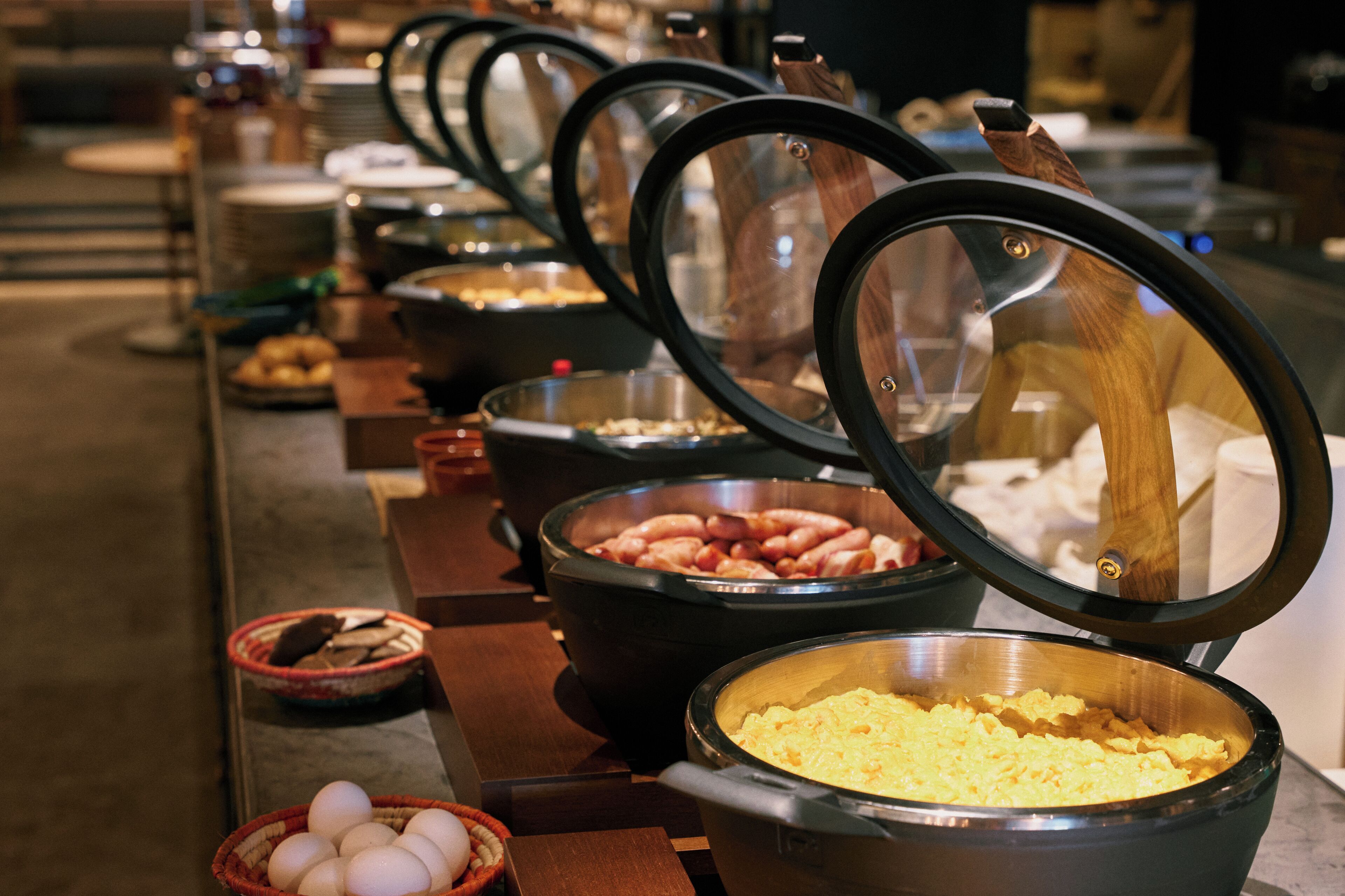 Una mesa de buffet con distintos alimentos, entre ellos huevos, salchichas y huevos revueltos.