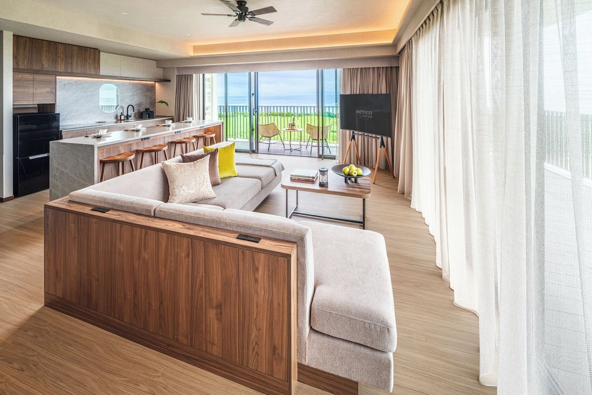 A modern living room with a sofa, a coffee table, and a TV. There is a balcony with a view of the ocean.