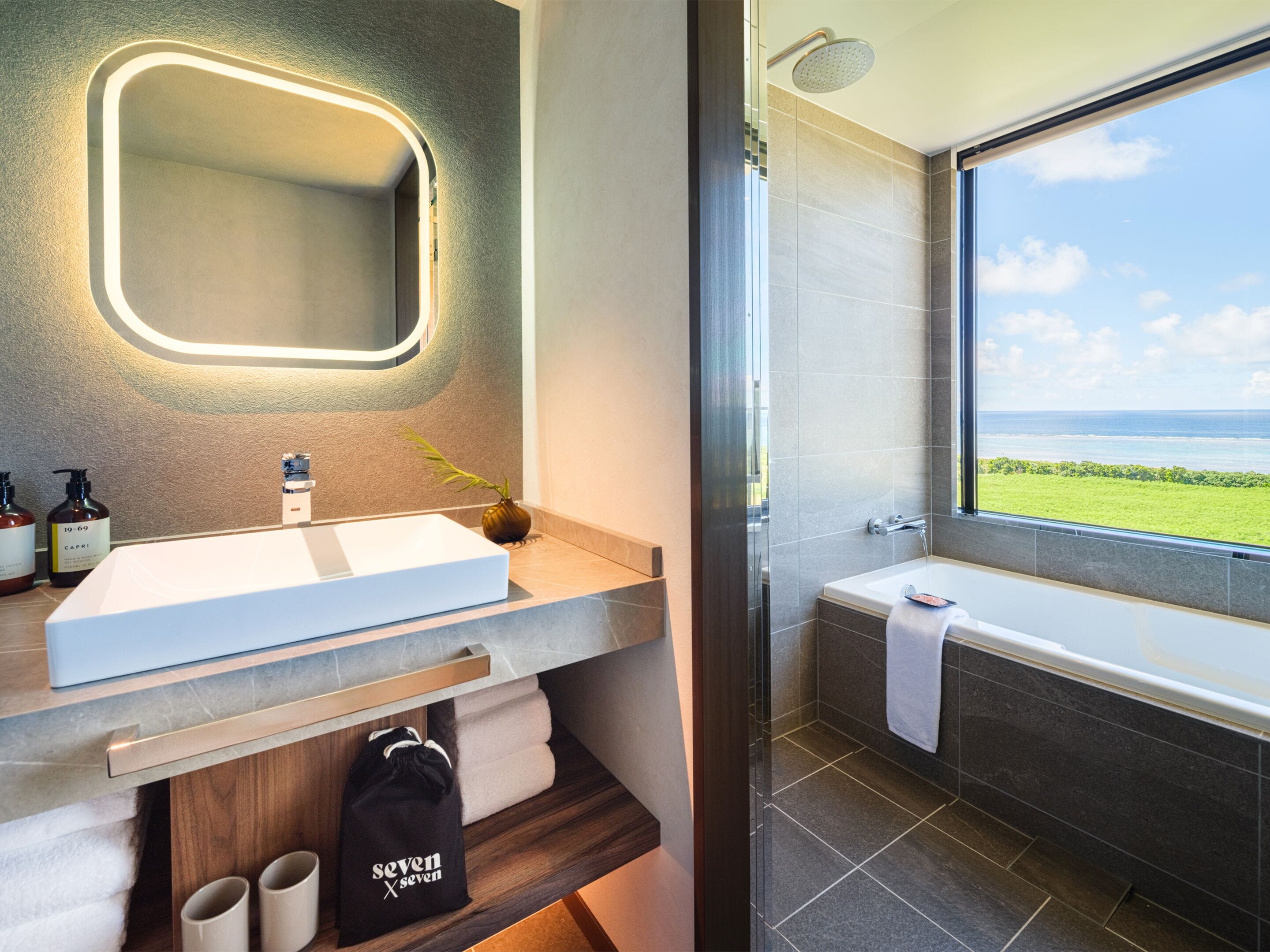 A modern bathroom with a large mirror, a white sink, and a bathtub. There is a window offering a view of the ocean.
