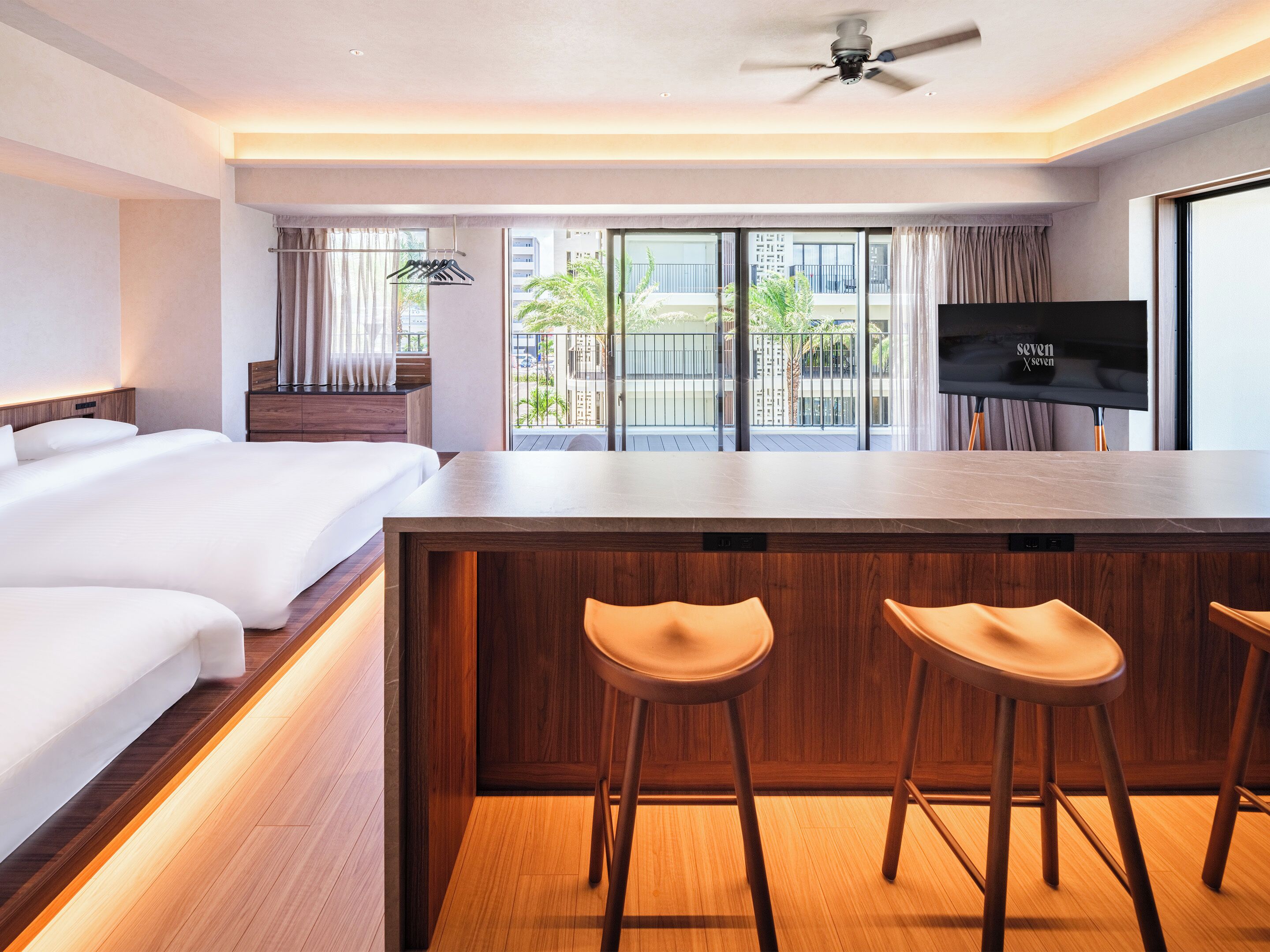 A hotel room with a large bed, a wooden bar area with stools, a flat-screen TV, and a balcony with a view of the city.