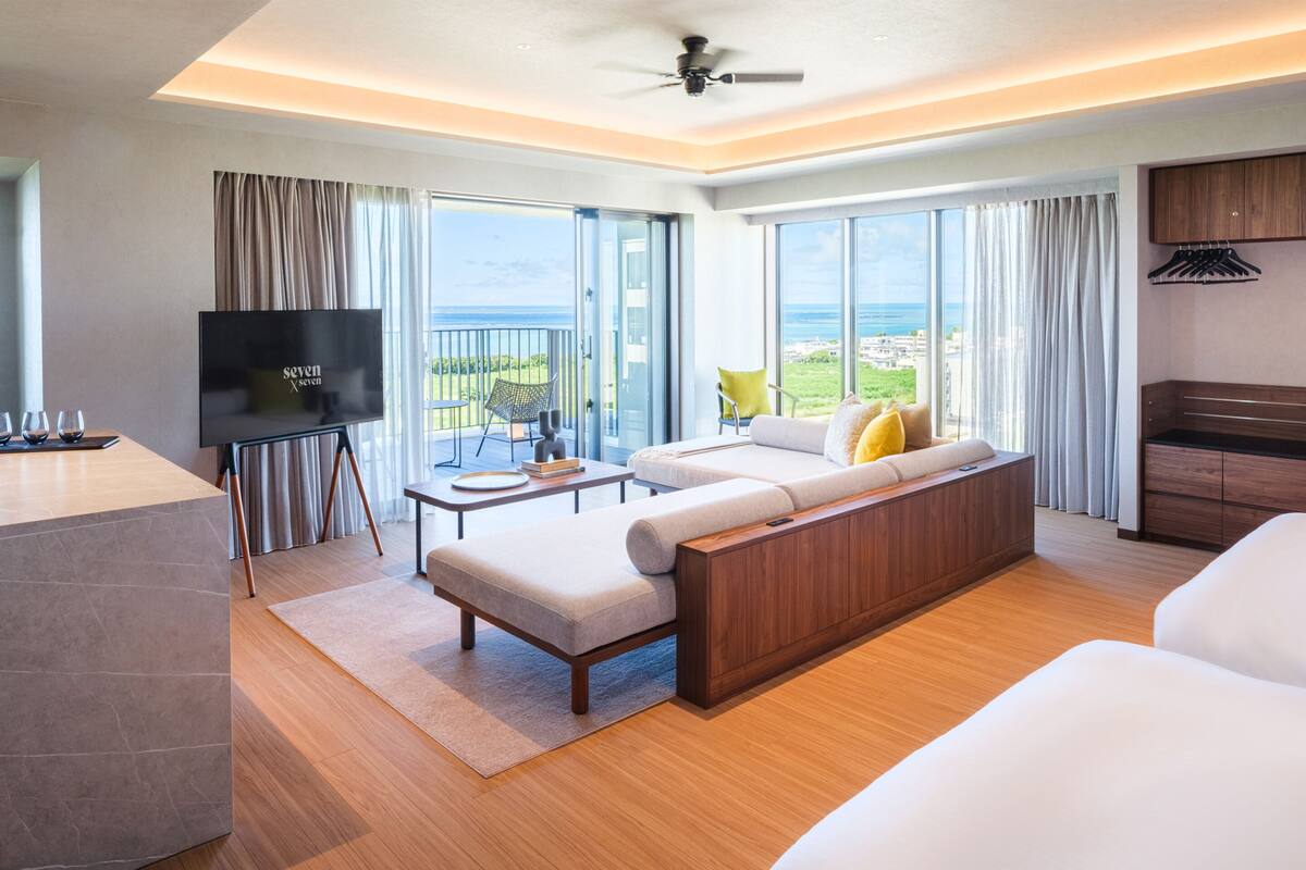 A modern hotel room with a large sofa, a flat-screen TV, a dining table, and a balcony with a view of the ocean.