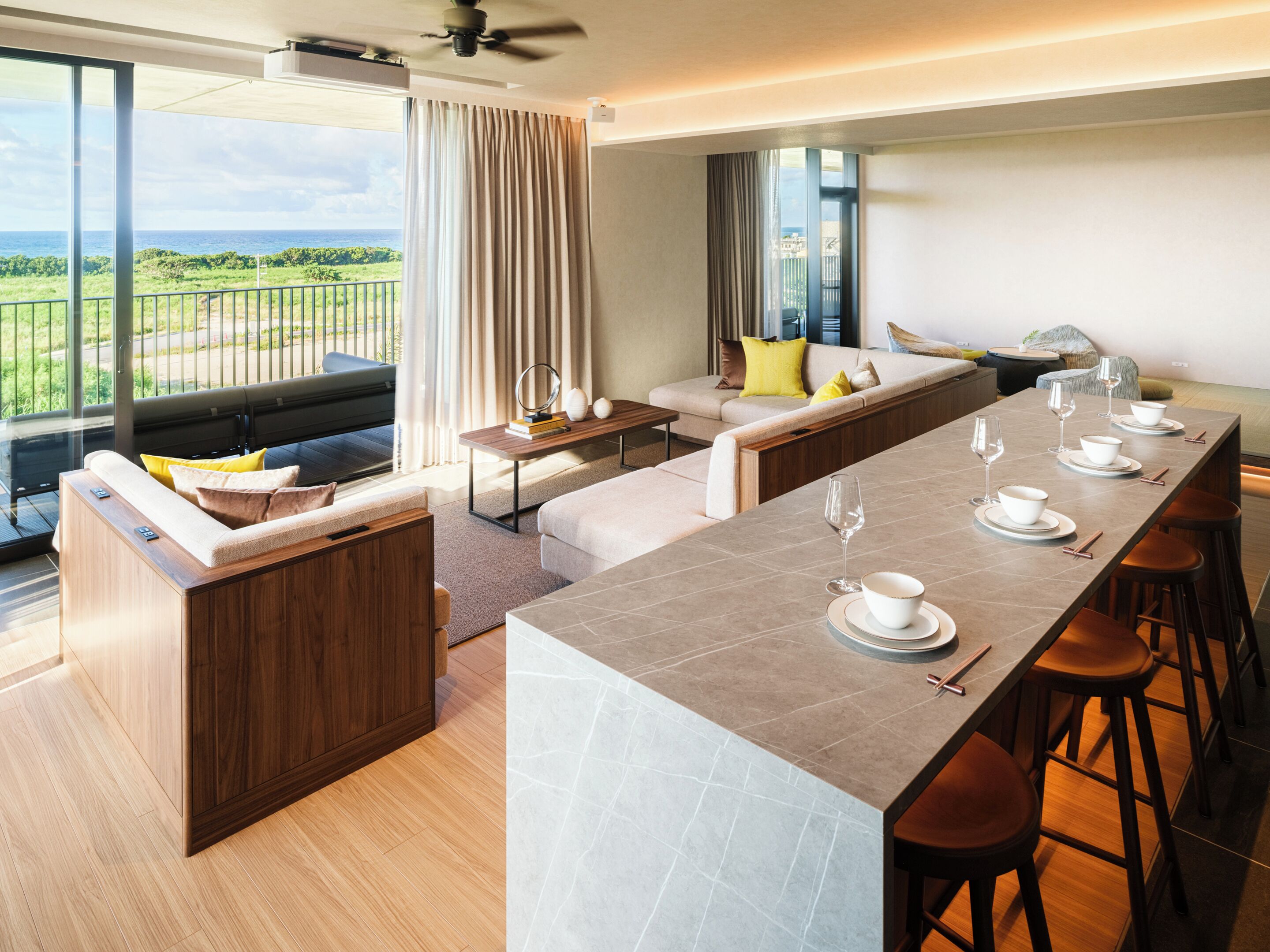A modern hotel room with a long dining table set for six, a sofa, a coffee table, and a balcony with a view of the ocean.
