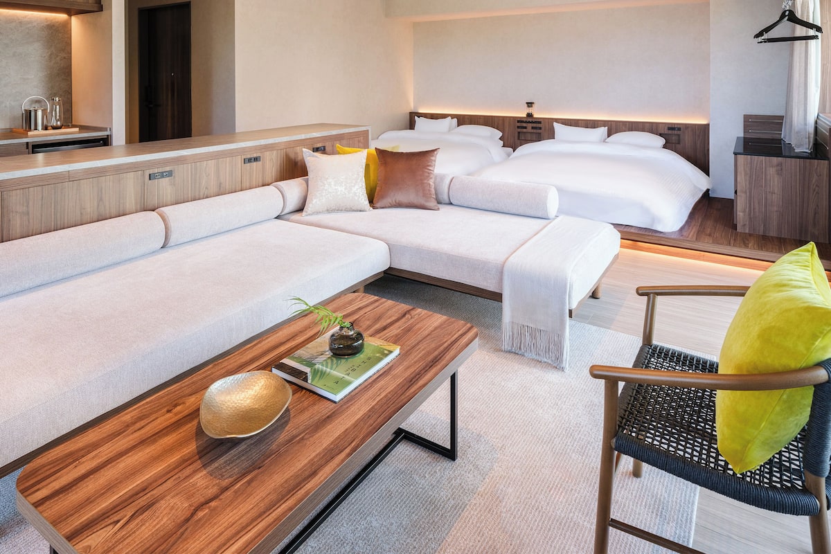 A modern hotel room with a sectional sofa, a single bed, a wooden coffee table with a book, a small table with a vase, and a chair with a yellow pillow.