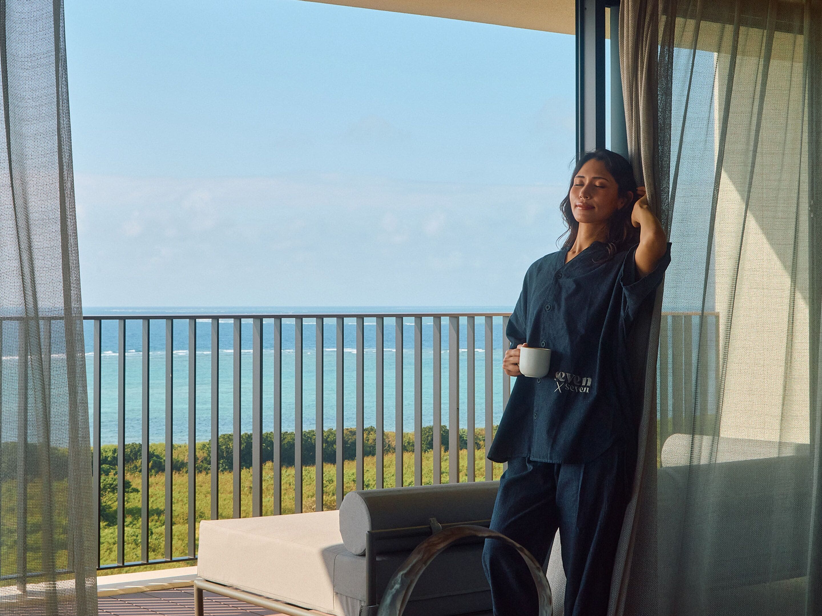 A balcony with a view of the ocean, a sofa, and a person holding a mug.