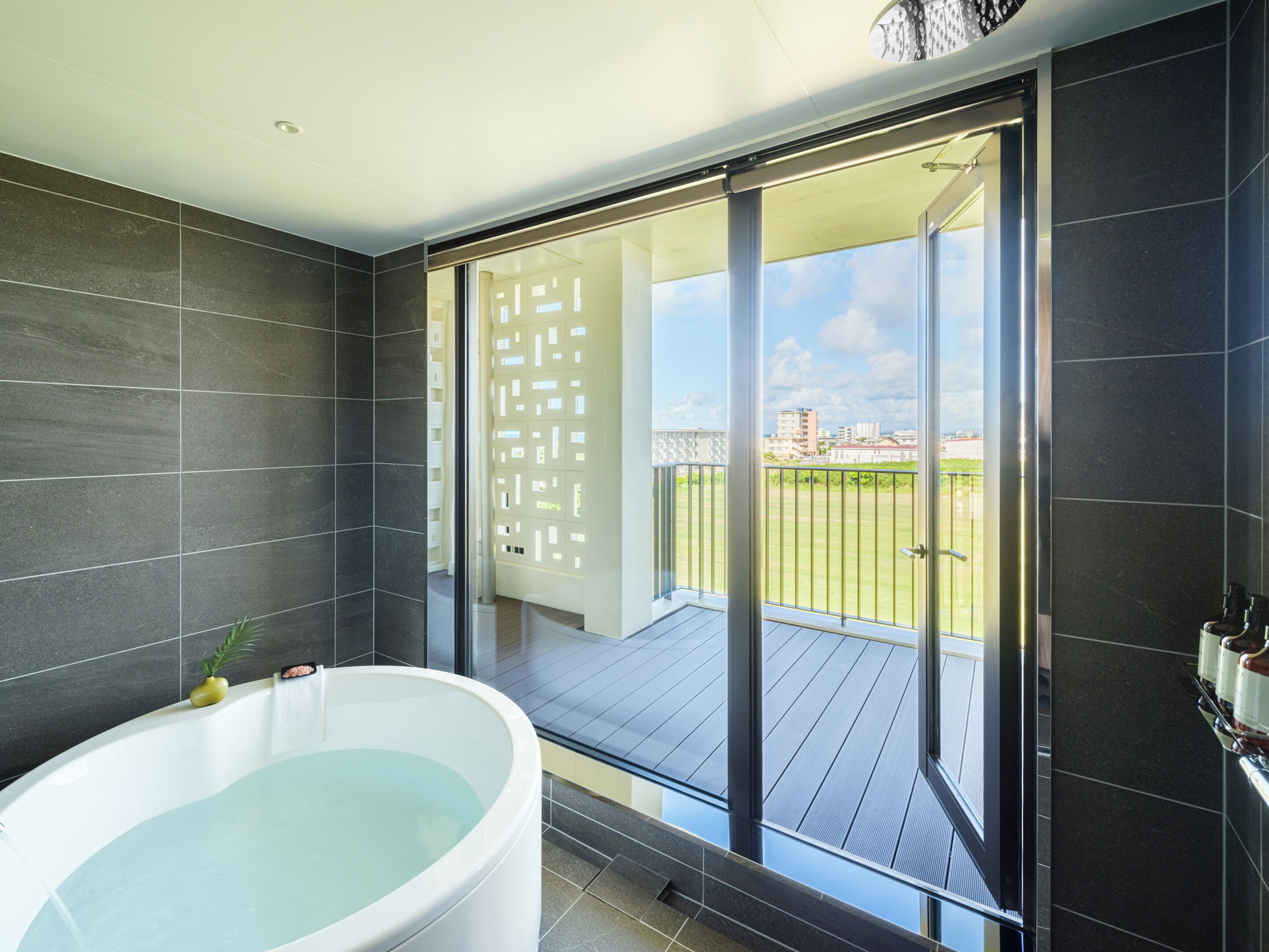 Un bagno moderno con una vasca da bagno ampia, pareti piastrellate scure e una porta scorrevole in vetro che dà su un balcone con vista su edifici e una zona verde.
