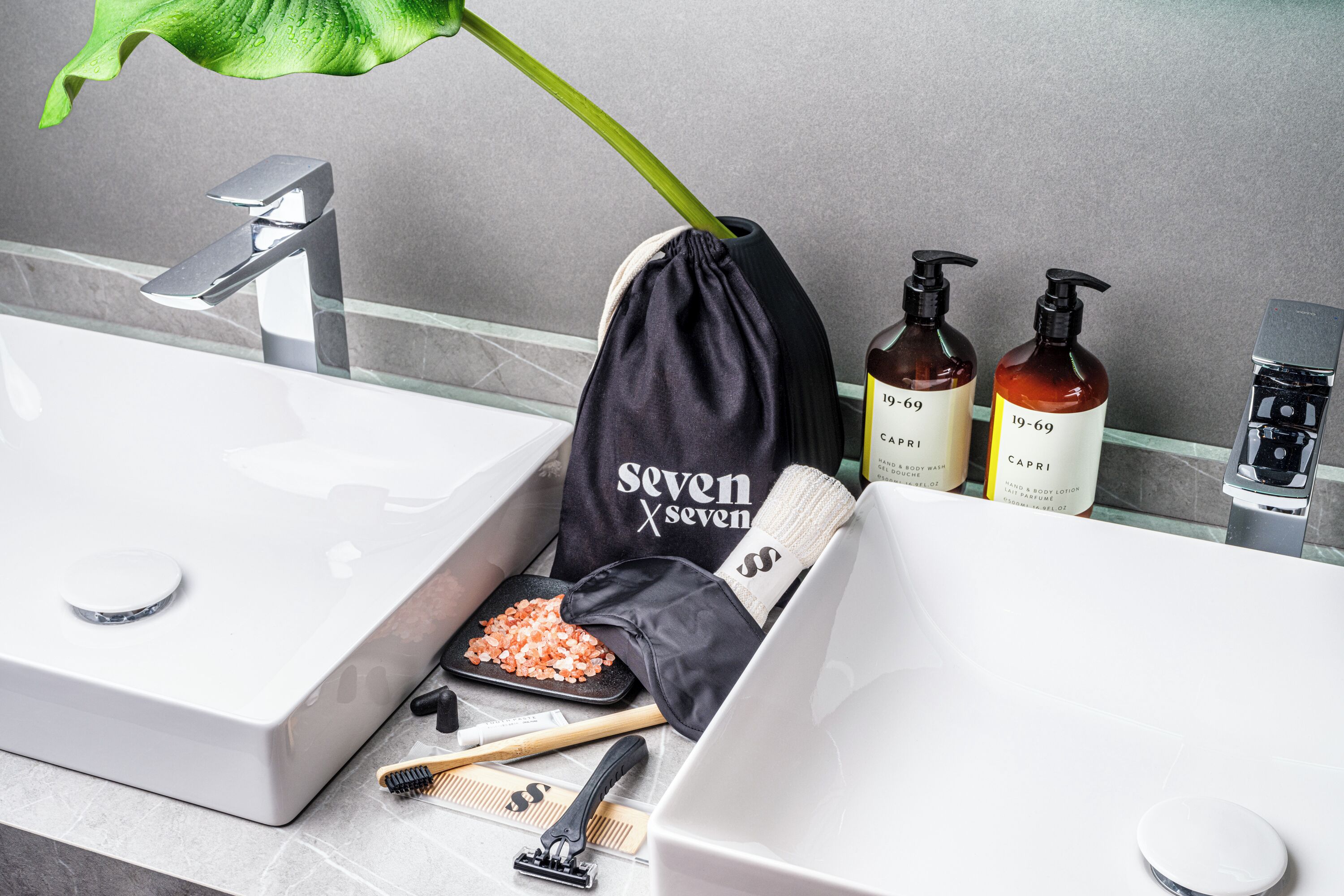 A bathroom with a white sink, a mirror, and toiletries including a black drawstring bag, two bottles of CAPRI body wash, and a green plant.