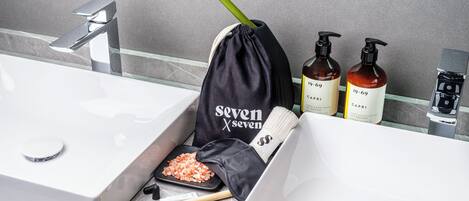 Un baño con un lavabo blanco, un espejo y artículos de aseo que incluyen una bolsa de estambre negra, dos botellas de CAPRI gel de ducha y una planta verde.